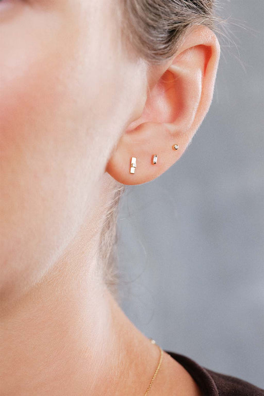 Close-up of model’s ear wearing three Sansoeurs earrings: a 18k gold Plaque Tag diamond baby earring, above it a 18k gold Baguette diamond baby earring, and higher an 18k gold Eye diamond baby earring.”